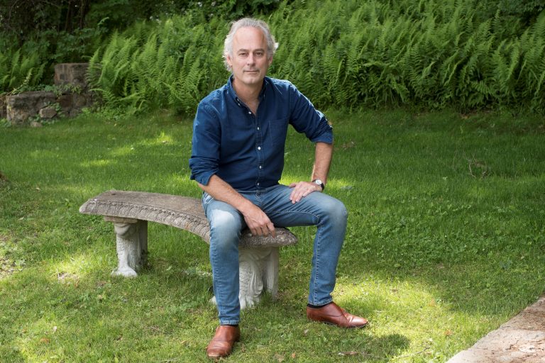 Amor Towles posa para a foto sentado em um banco de pedra, em um jardim