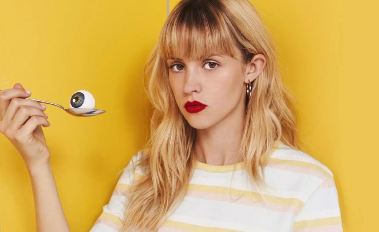 Angèle posa para a foto segurando uma colher com um olho em cima, em fundo amarelo