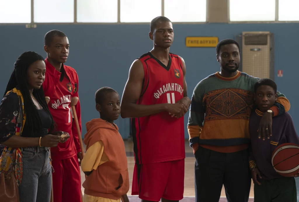 Seis pessoas negras estão em pé. Cinco homens, sendo duas crianças, e uma mulher. Dois deles vestem um uniforme vermelho de basquete e uma das crianças segura uma bola de basquete.