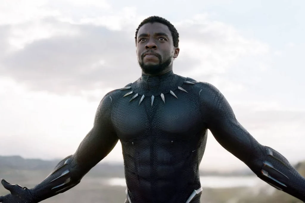 O ator Chadwick Boseman, negro, em Pantera Negra, de braços abertos vestindo o traje do super-herói.