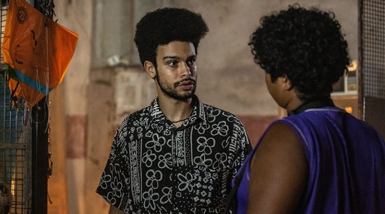O ator Sérgio Malheiros, negro, olha para um homem, também negro, que está de costas.