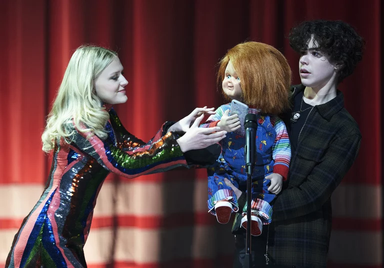 Jake (Zacakary Arthur), jovem branco e moreno, e Lexy (Alyvia Alyn Lind), jovem branca e loira, estão em cima de um palco. Jake segura Chucky e Lexy tenta segurá-lo.