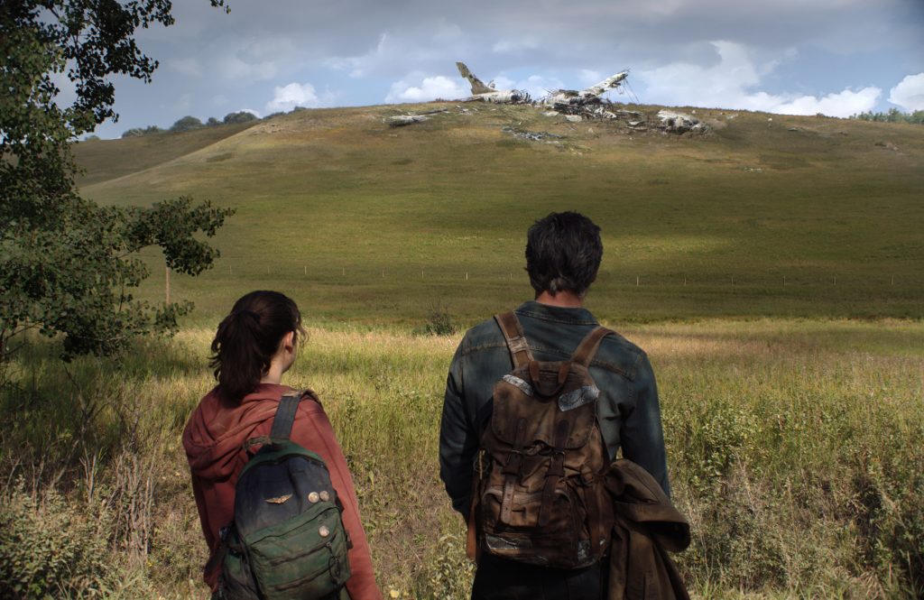 Pedro Pascal e Bella Ramsey aparecem de costas e ao fundo uma paisagem verde o céu azulado na parte de cima da imagem, em cena em The Last of Us.