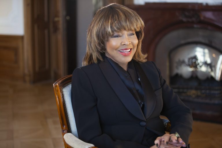 Tina Turner em uma entrevista do documentário TINA. Ela usa roupa preta e esta sentada em uma cadeira, em uma sala clássica, com piso de madeira e lareira ao fundo